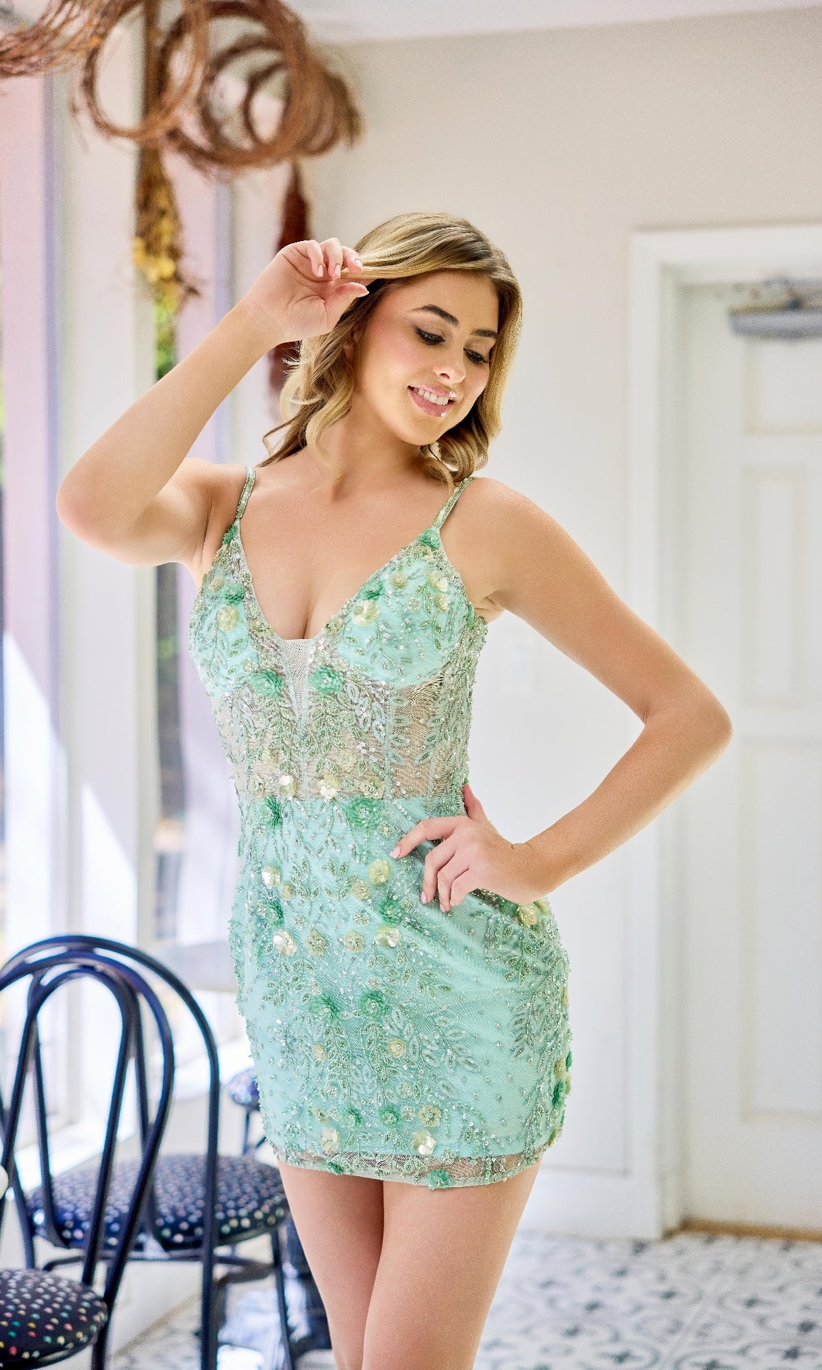 A woman smiles and poses indoors in the Primavera 4402, a light green floral-beaded short hoco dress, with one hand gracefully raised near her head.